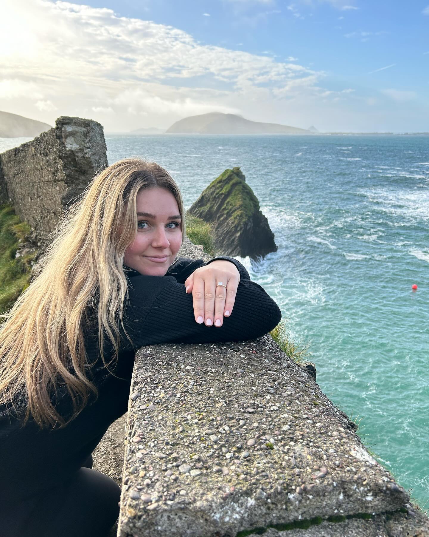 Caitlin leaning on a seaside wall, smiling with wind in her hair.